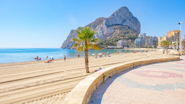 A beach with a curved walkway, palm trees, and a mountain in the background. - Apartment Turmalina - PlusHolidays (Calpe)
