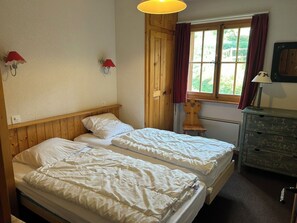 Quarto com duas camas, um guarda-roupa de madeira, uma janela com cortinas e uma luminária fixada na parede.