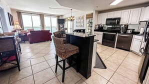 A modern kitchen with white cabinets, a black countertop, and stainless steel appliances. There is a dining area with a red sofa and wicker chairs. - PARADISE ON THE OCEAN! TWO FREE BEACH CHAIR, FREE VIP PARKING SPACE! (PANAMA CITY BEACH)