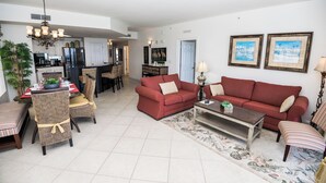 A spacious living room with a red sofa, a dining area with wicker chairs, and a kitchen in the background. - PARADISE ON THE OCEAN! TWO FREE BEACH CHAIR, FREE VIP PARKING SPACE! (PANAMA CITY BEACH)