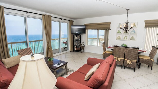 A spacious living room with a red sofa, a dining area with wicker chairs, a flat-screen TV, and a balcony with a view of the ocean. - PARADISE ON THE OCEAN! TWO FREE BEACH CHAIR, FREE VIP PARKING SPACE! (PANAMA CITY BEACH)