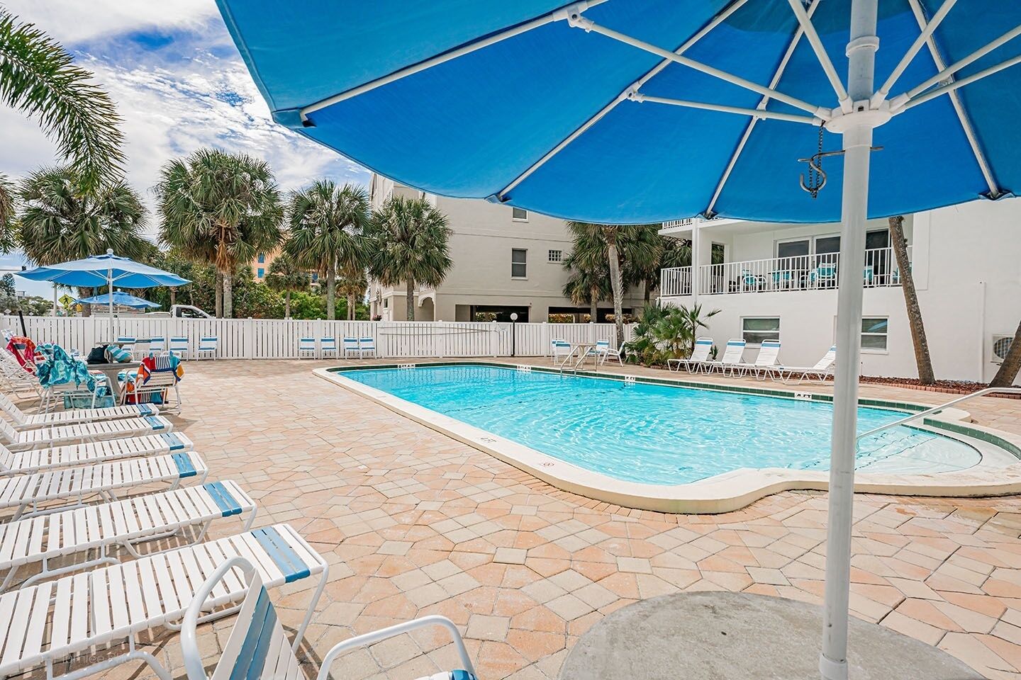 Area piscina con poltrona a sdraio e ombrelloni, circondata da palme e un edificio con balconi.