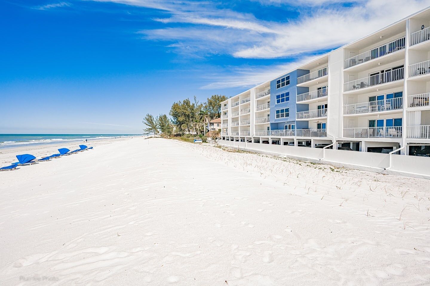 A beachfront hotel with multiple floors, balconies, and a sandy beach with umbrellas.