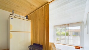 Ein kleines Zimmer mit einem weißen Kühlschrank, einem kleinen schwarzen Hocker, einer Holzbalkendecke und einem großen Fenster mit Blick nach draußen.
