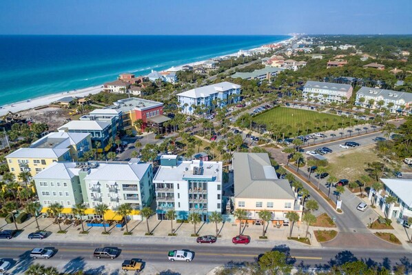 An aerial view of a coastal town with a beachfront, multi-story buildings, and a road with parked cars.