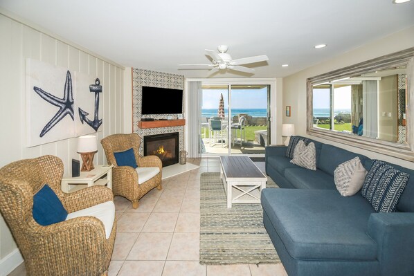 A living room with a fireplace, a television, wicker chairs, a blue sofa, and a view of the ocean. - Beachfront lower cottage, panoramic view, D31 North Coast Village (Oceanside)