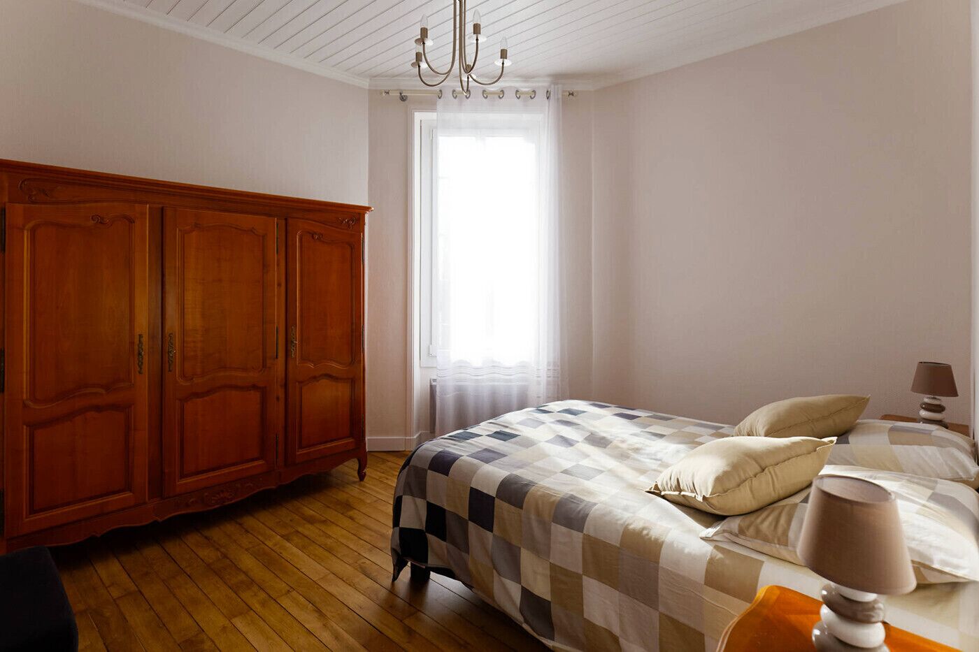 Un dormitorio con un armario de madera, una cama con edredón a cuadros y una ventana con cortinas de gasa.