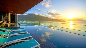 Une piscine à débordement avec des garde-corps en verre transparent, un plafond en bois et une vue imprenable sur le coucher de soleil sur l’océan.