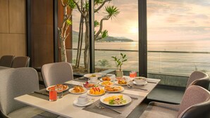 Une table à manger dressée avec différents plats, un verre de jus d’orange et une vue sur l’océan par de grandes fenêtres.