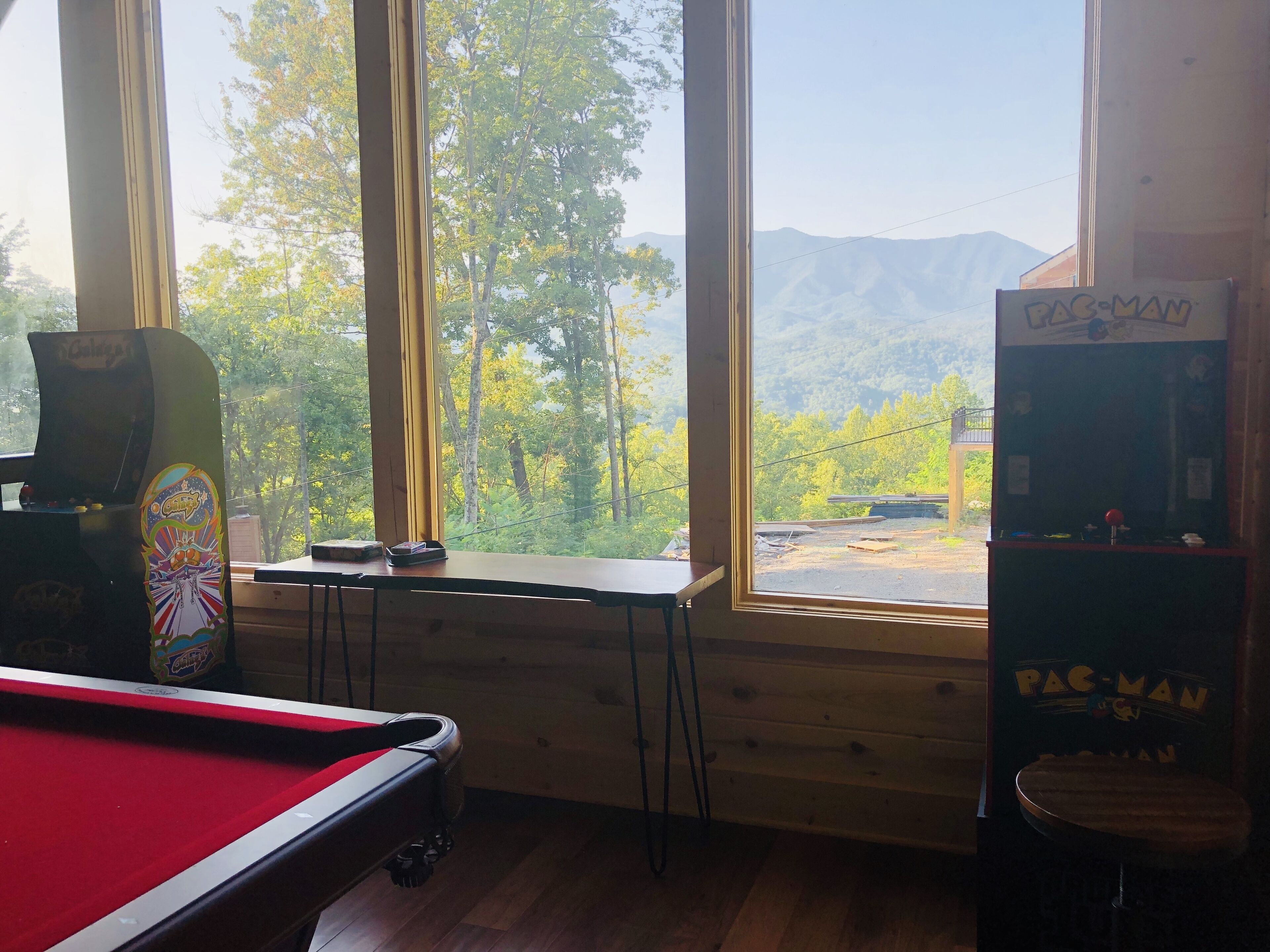 A room with a pool table, a Pac-Man arcade machine, and a view of a forested area through large windows.