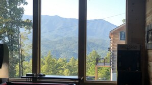 Una mesa de billar en una habitación con ventanas amplias que dan a una cadena montañosa boscosa.