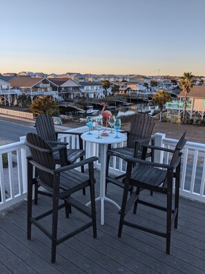 Una terraza con cuatro sillas Adirondack y una mesa redonda, con vista a un puerto deportivo con barcos y casas.