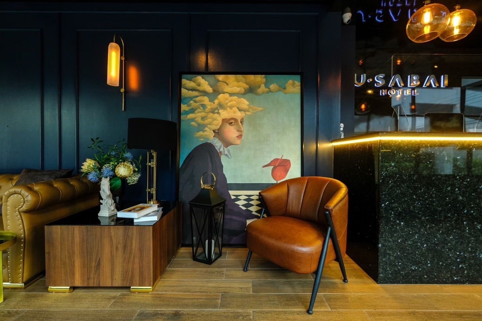 A reception area with a wooden desk, a leather armchair, a painting of a woman, and a sign for 'U.SABAI HOTEL'.