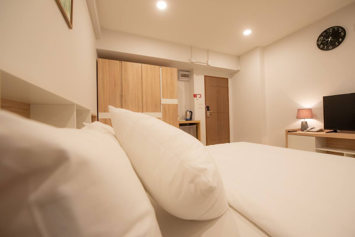 Quarto de hotel com uma cama, guarda-roupa de madeira, televisão e relógio na parede.