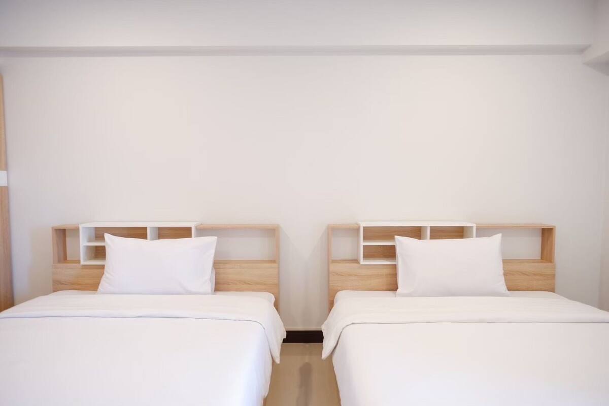 Duas camas de solteiro com cabeceiras de madeira e roupa de cama branca em um quarto com paredes brancas sem decoração.