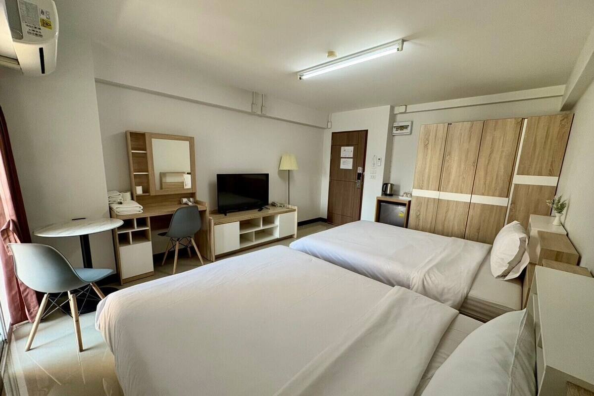 Quarto de hotel com duas camas, uma escrivaninha com computador, televisão, pequeno espaço de cozinha e guarda-roupa.
