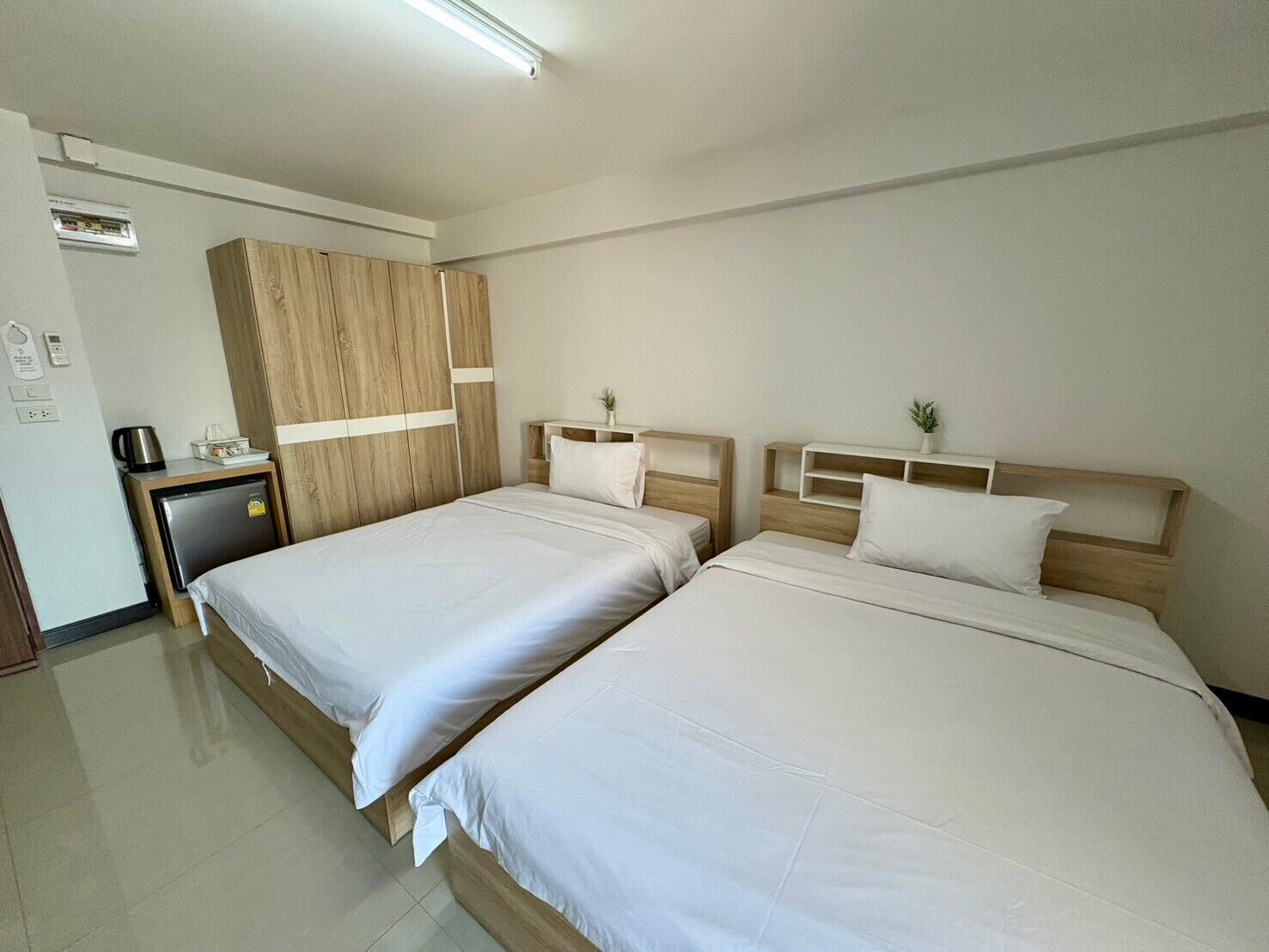 A hotel room with two beds, a wooden wardrobe, a small desk with a kettle, and a wall-mounted air conditioner.