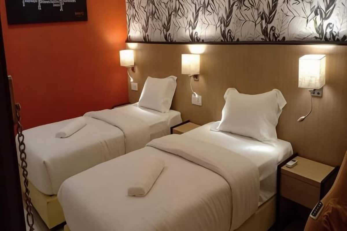 A hotel room with two beds, each with a pillow and a blanket, bedside lamps, and a nightstand with a remote control.