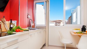 A modern kitchen with a red accent wall, a window with a city view, and a dining area with a white chair. - A. Corales del Mar in Alcudia Beach (Port d´Alcudia)