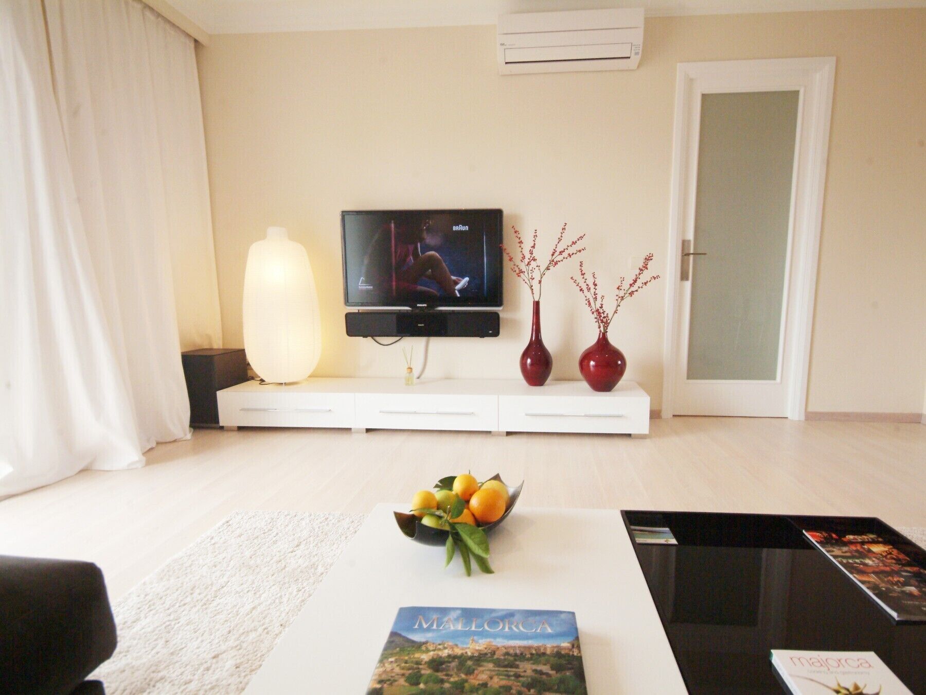 A modern living room with a flat-screen TV, a white sofa, a coffee table with a fruit bowl, and a magazine.