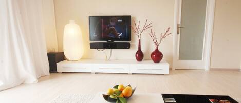 A modern living room with a flat-screen TV, a white sofa, a coffee table with a fruit bowl, and a magazine.