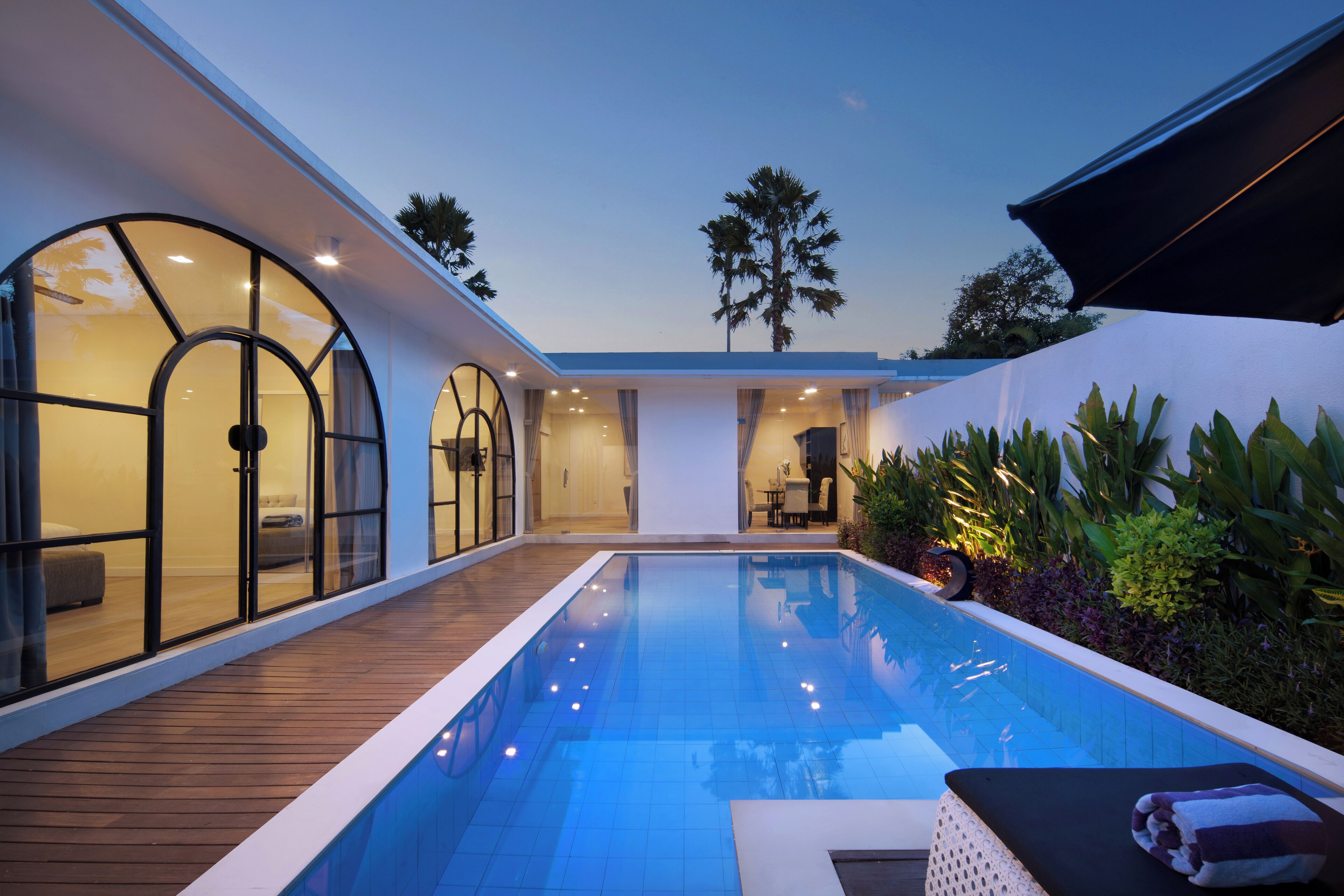 A modern poolside area with a wooden deck, a pool, and a lounge area with a sunbed and a towel.
