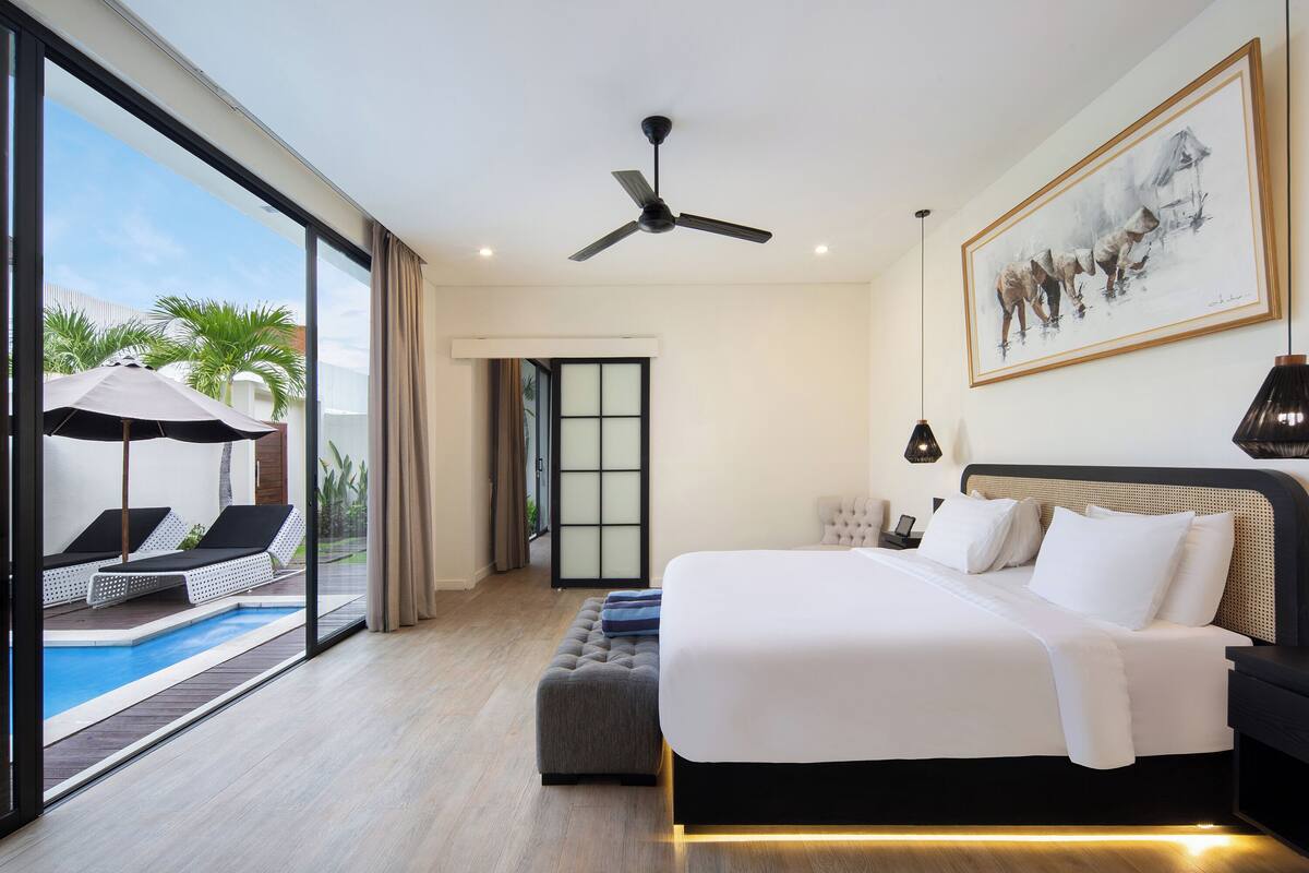 Quarto de hotel moderno com uma cama grande, um ventilador de teto, um quadro na parede e vista para uma piscina e palmeiras do lado de fora.