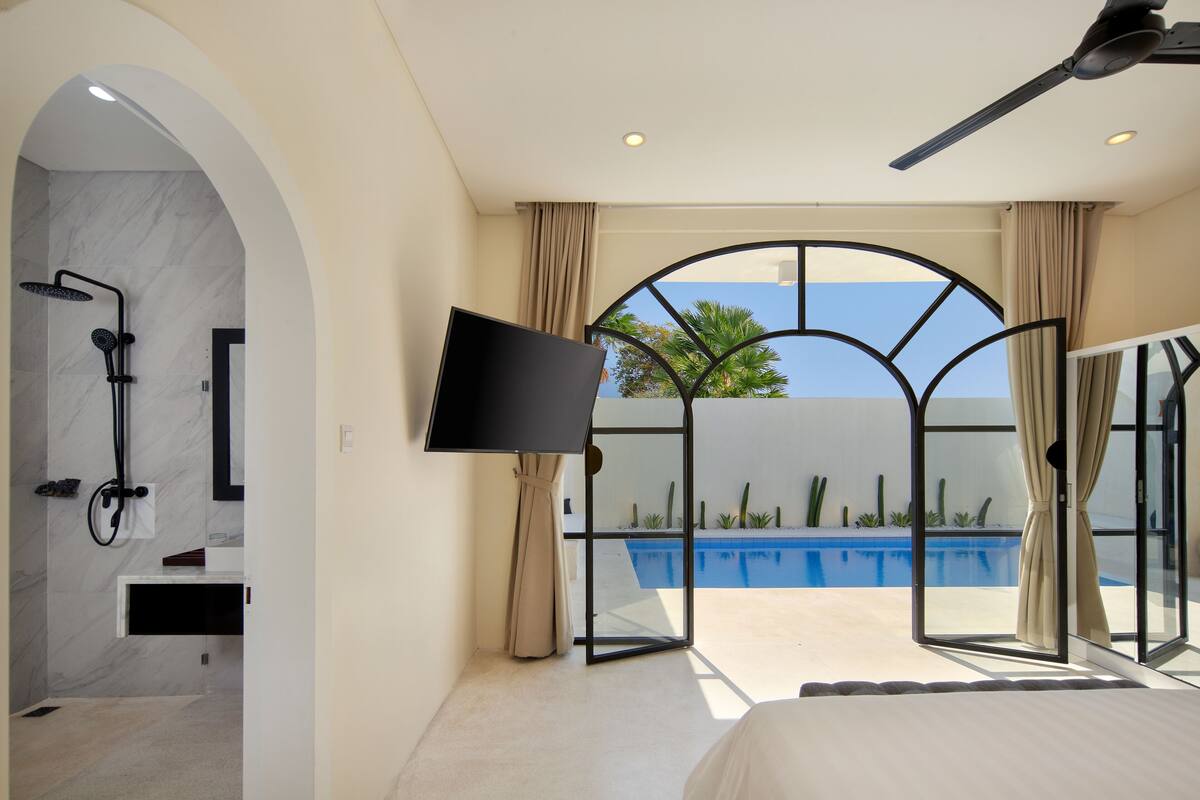 Quarto de hotel moderno com cama grande, televisão, chuveiro e vista para uma piscina.