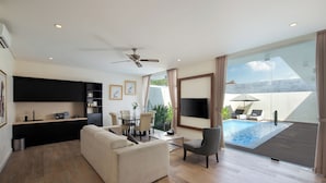 A modern living room with a sofa, a dining table, and a TV. There's a pool area with a chair and a table. - The Grand Daha Luxury Villas (Seminyak)