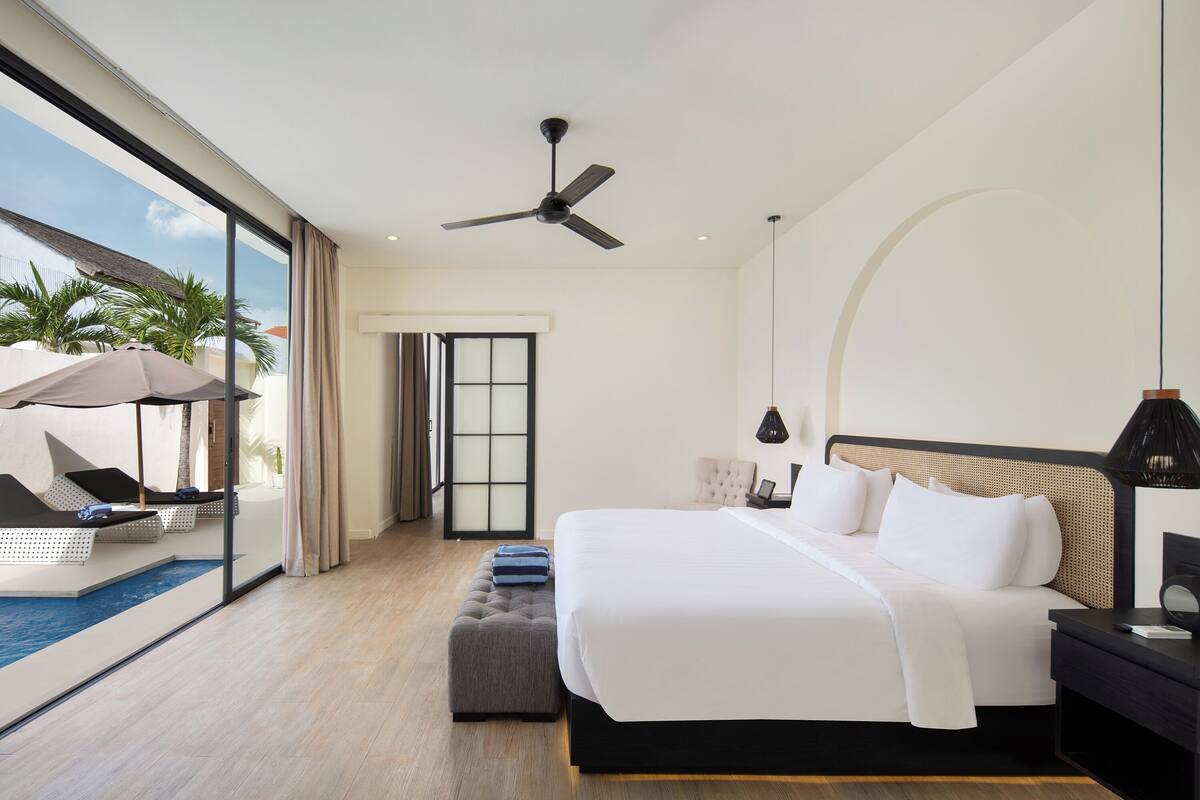 Quarto de hotel moderno com uma cama grande, ventoinha no teto, vista para a piscina e uma varanda com cadeiras de descanso.