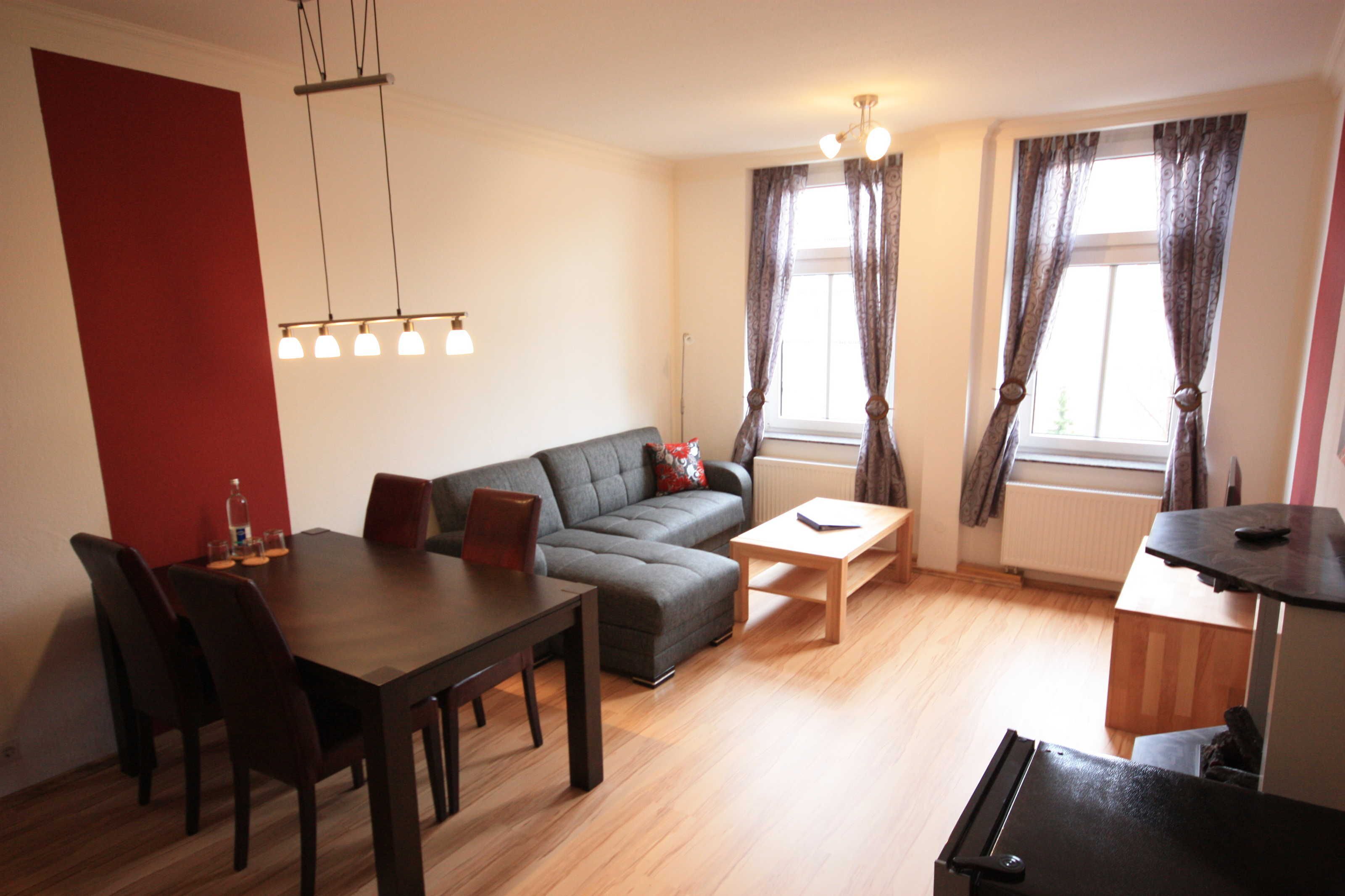 Ein modernes Wohnzimmer mit Esstisch, Sofa und Couchtisch. Ein Heizkörper, eine Lampe und ein Fenster mit Vorhängen sind ebenfalls vorhanden.