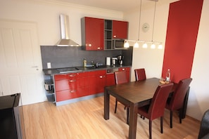 Cocina compacta con armarios rojos, microondas y cafetera. Hay una mesa de comedor con sillas y un reloj en la pared.