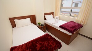 A hotel room with two beds, a window with curtains, and a small bedside table with a phone and a plant. - Hotel Kashi Khand (Varanasi)