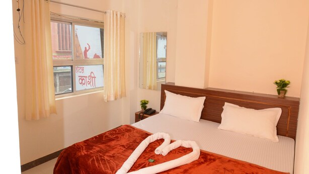 A neatly made bed with a red blanket and white pillows, a ceiling fan, and a window with curtains. - Hotel Kashi Khand (Varanasi)