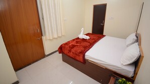 A hotel room with a wooden door, a bed with a red bedspread, white pillows, and a small table with a plant. - Hotel Kashi Khand (Varanasi)