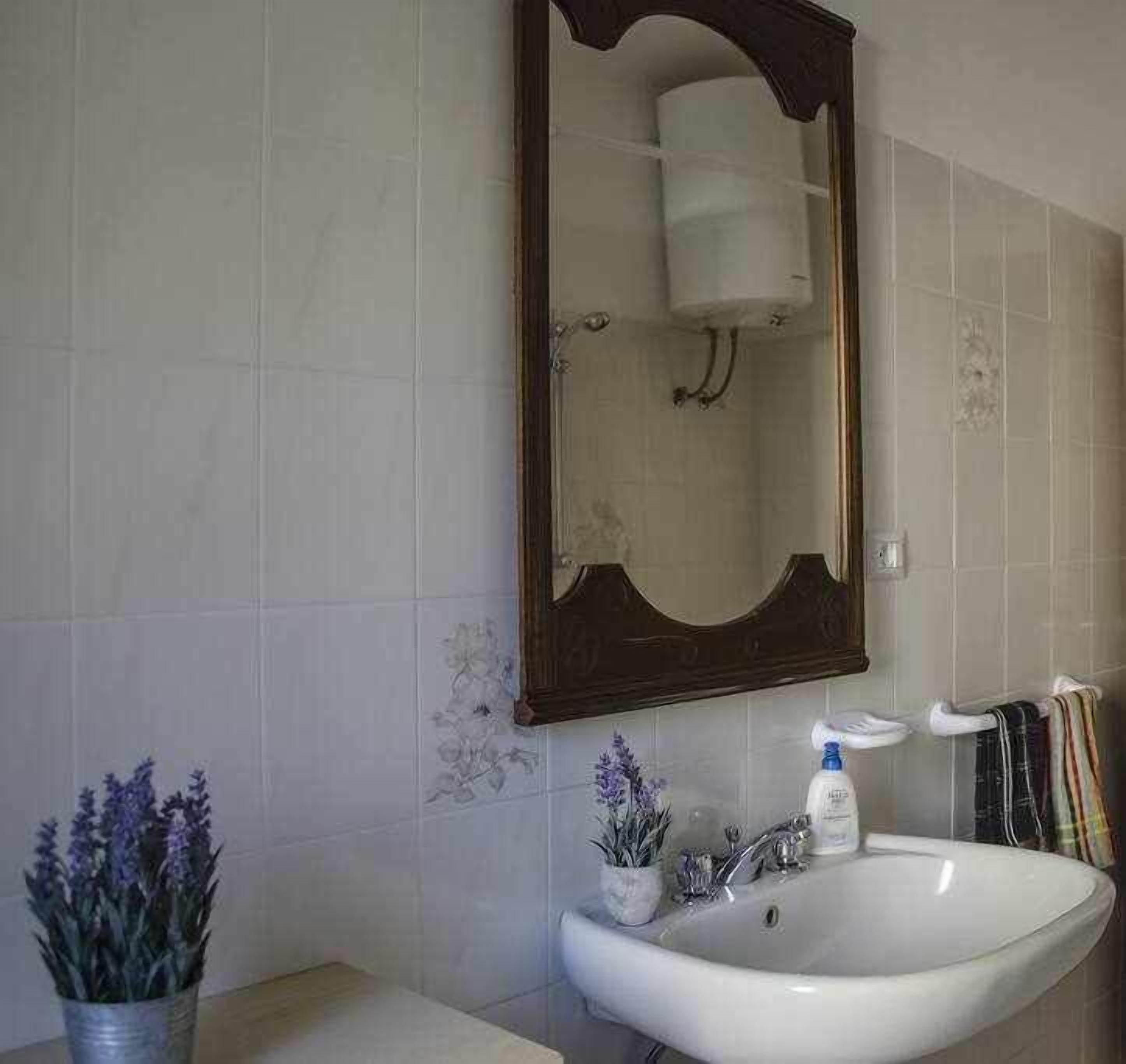 Un bagno con un lavandino bianco, uno specchio e un vaso con fiori viola.