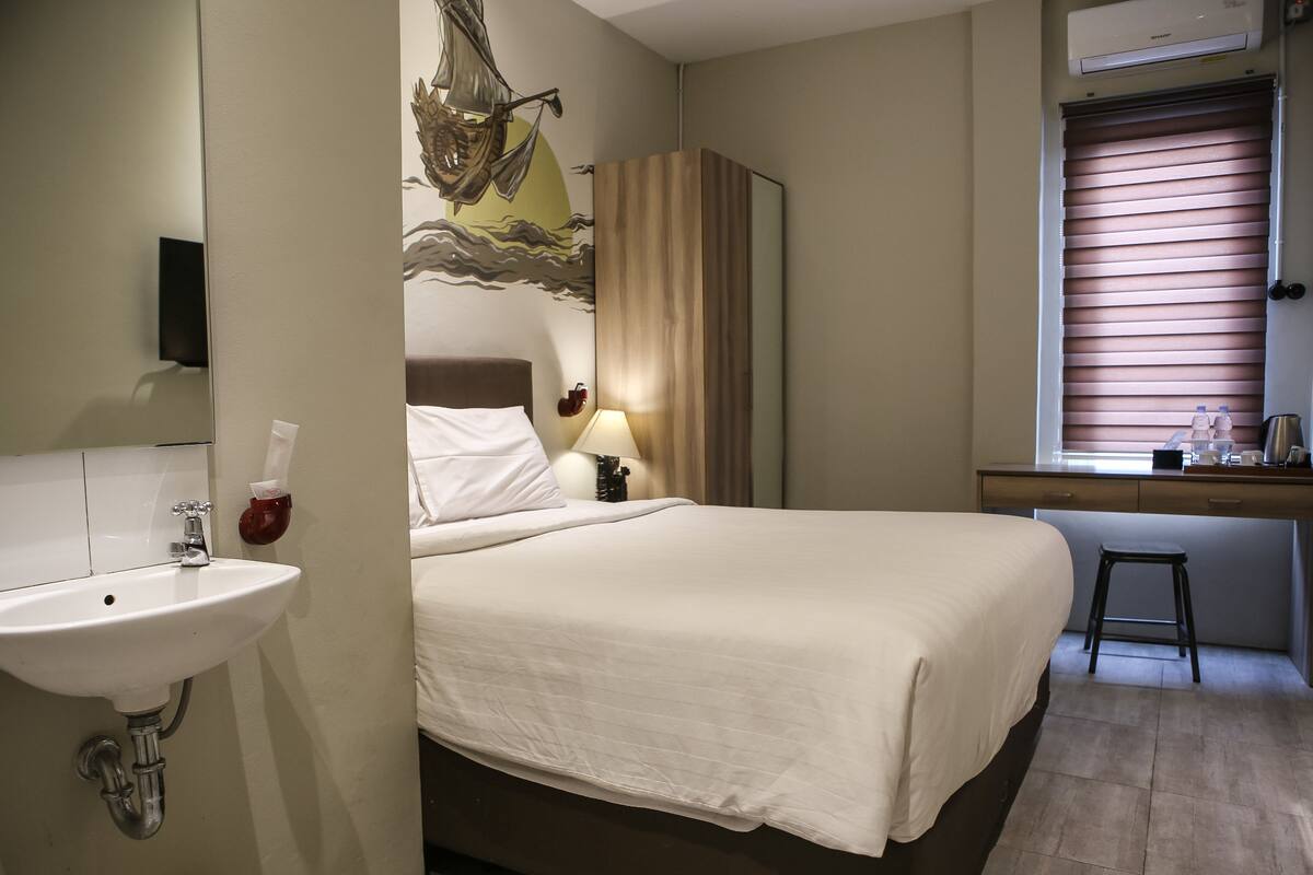 A hotel room with a bed, a bedside table with a lamp, a desk with a computer, a window with blinds, and a bathroom with a sink and mirror.