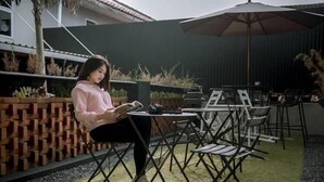 Una persona sentada a una mesa con un libro, al aire libre, con un área de patio, sillas y una mesita.