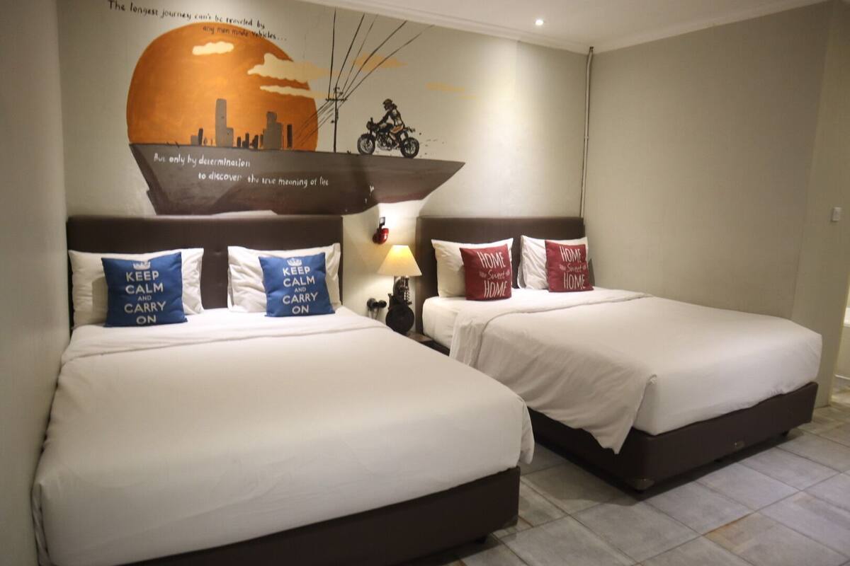 A hotel room with two beds, each with a pillow that says 'KEEP CALM AND CARRY ON,' a wall mural with a sunset and a motorcycle, and a nightstand with a lamp.