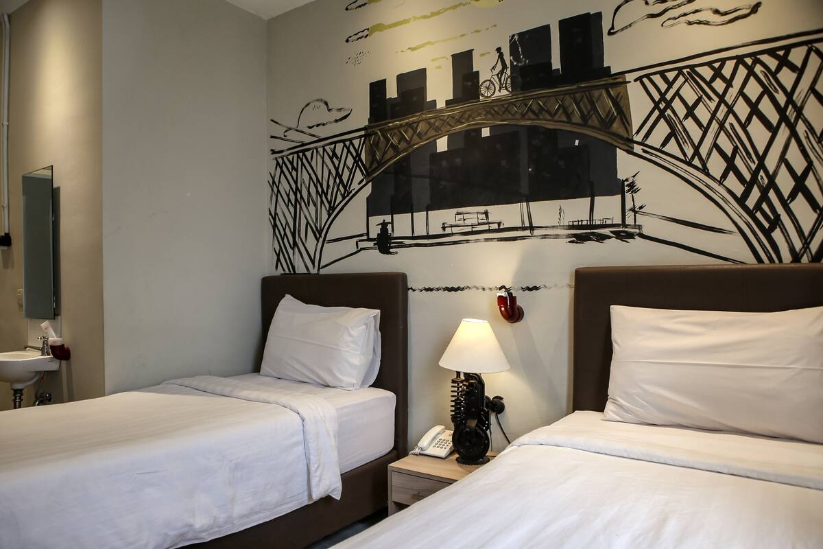 A hotel room with two single beds, a bedside table with a lamp, a phone, and a wall mural depicting a bridge and cityscape.