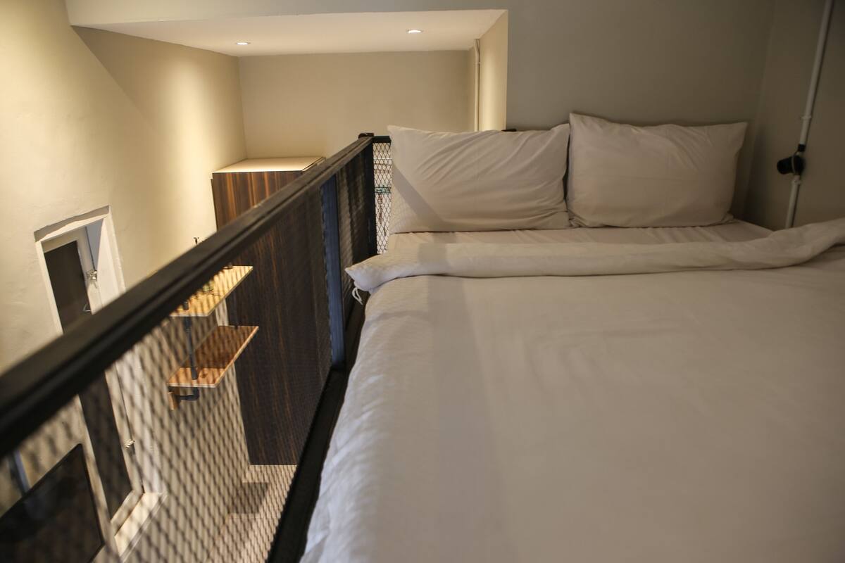 A hotel room with a bed, a wooden headboard, a small wooden shelf, and a window with a view of the outside.