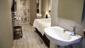 Habitación de hotel con una cama, un lavamanos blanco, un espejo y un taburete pequeño.