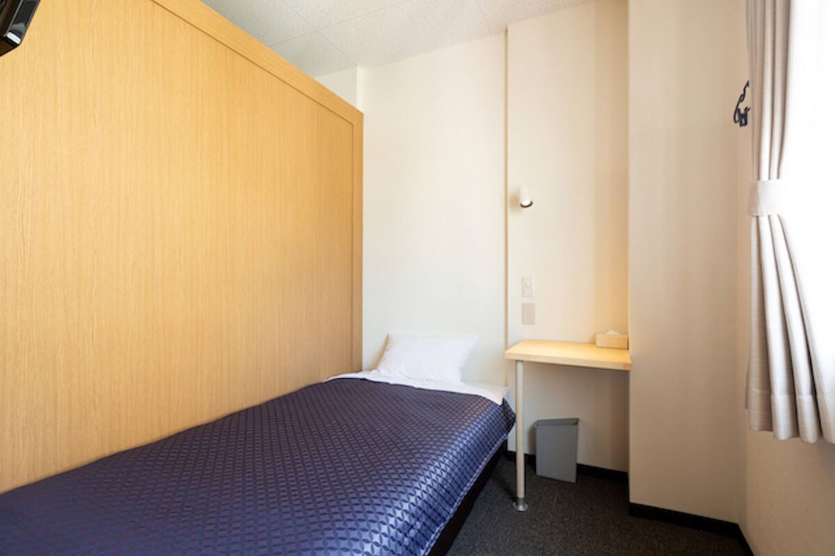 A small, single-bed room with a wooden headboard, a bedside table, and a small trash bin.