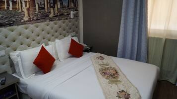 Quarto de hotel com uma cama grande, duas almofadas vermelhas, um corredor floral e um mural de ruínas antigas na parede.