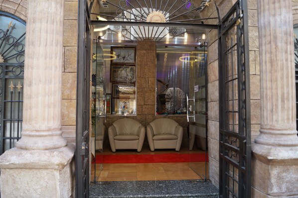 A hotel entrance with a red carpet leading to two leather chairs, a glass door with a metal gate, and a decorative ceiling fan.
