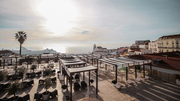 A rooftop terrace with outdoor seating, palm trees, and a view of the city and sea.