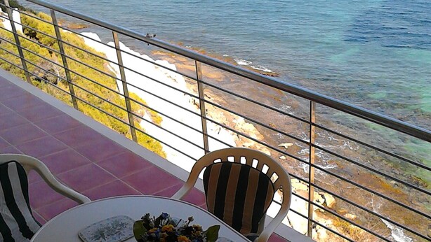 A round table with a vase of flowers, two chairs, and a view of the sea and rocky shore.