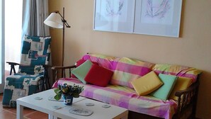A living room with a colorful sofa, a small white table with a vase, and two framed artworks on the wall.