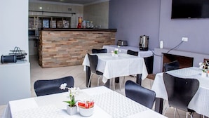 A dining area with tables and chairs, a kitchen in the background, and a flat-screen TV mounted on the wall. - Rose Villa Lodge (Kitwe)