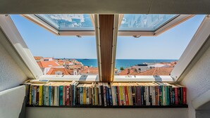 Ein Dachfenster mit Blick auf eine Küstenstadt und ein Bücherregal voller Bücher.